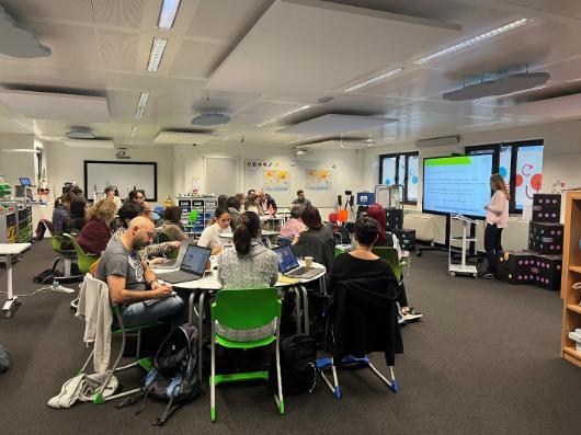 Teachers sitting in round tables working on their laptop while a person is presenting a ppt.