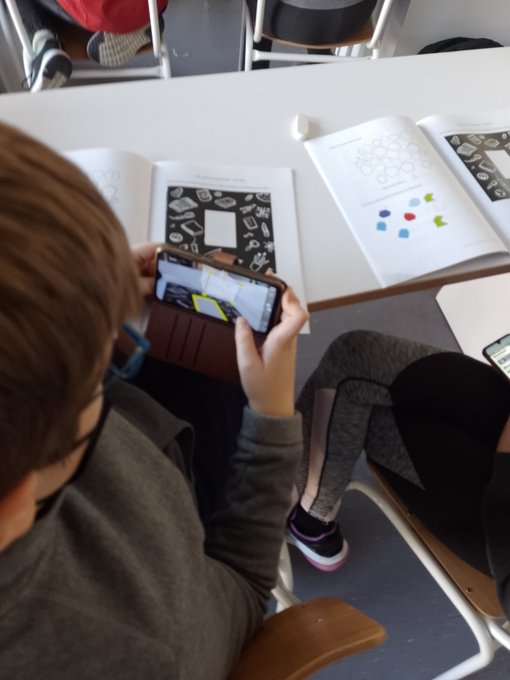 A child holding a phone where a 3D image of a cube can be seen on top of their book (which has no cube)